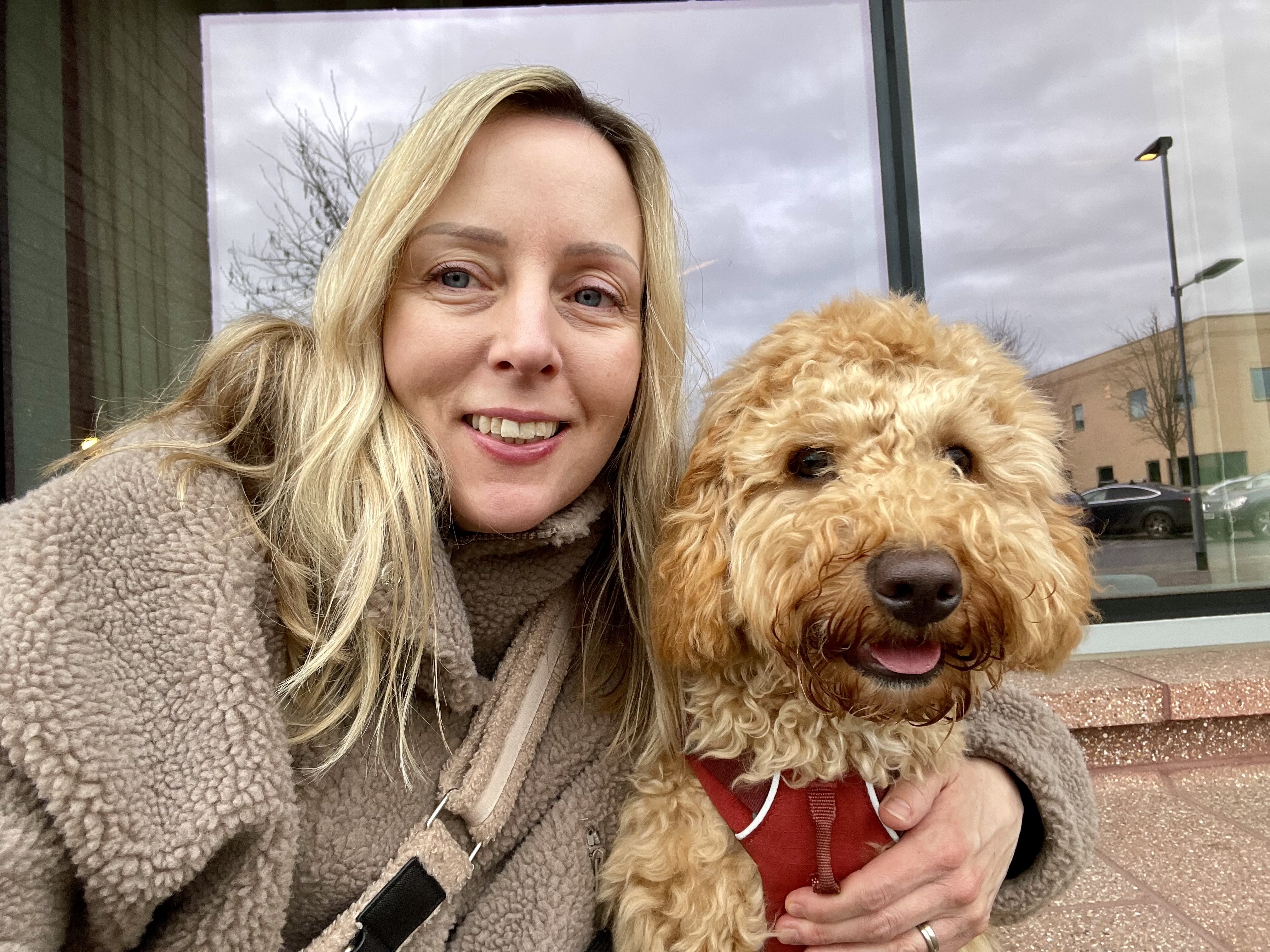 A woman with long blonde hair, wearing a fuzzy gray coat, poses for a selfie. Sitting closest to the woman is a small, cur...
